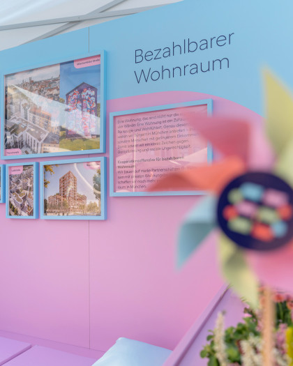 Blick auf das innere des Messestandes der Münchner Wohnen. An der bunten Wand hängen mehrere Bilder von Bauprojekten. Daneben steht "Bezahlbarer Wohnraum". Im Vordergrund erkennt man ein Windrad im Münchner Wohnen Design.
