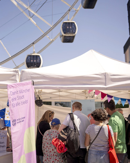 Blick auf den Stand der Münchner Wohnen mit einer Menschengruppe davor. Im Hintergrund sieht man die Gondeln eines Riesenrades.