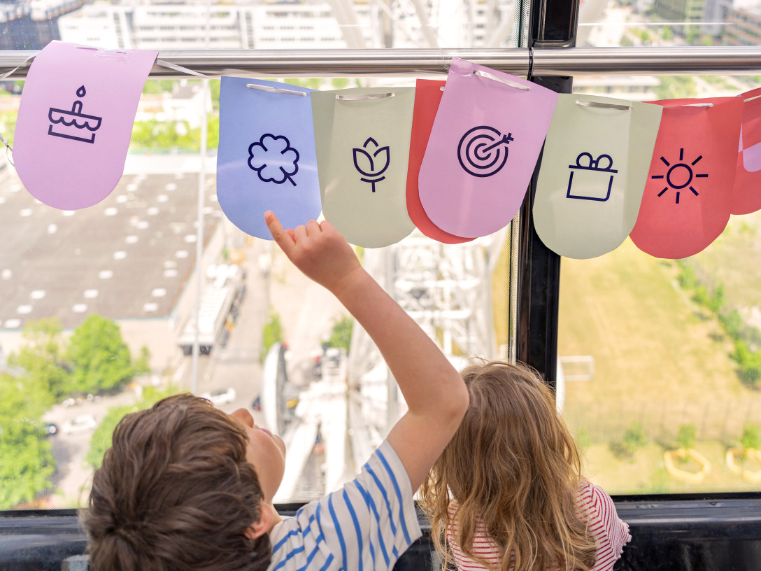 Zwei Kinder sitzen unter einer bunten Girlande im Münchner Wohnen Design, die verschiedene Symbole rund ums Wohnen zeigt.