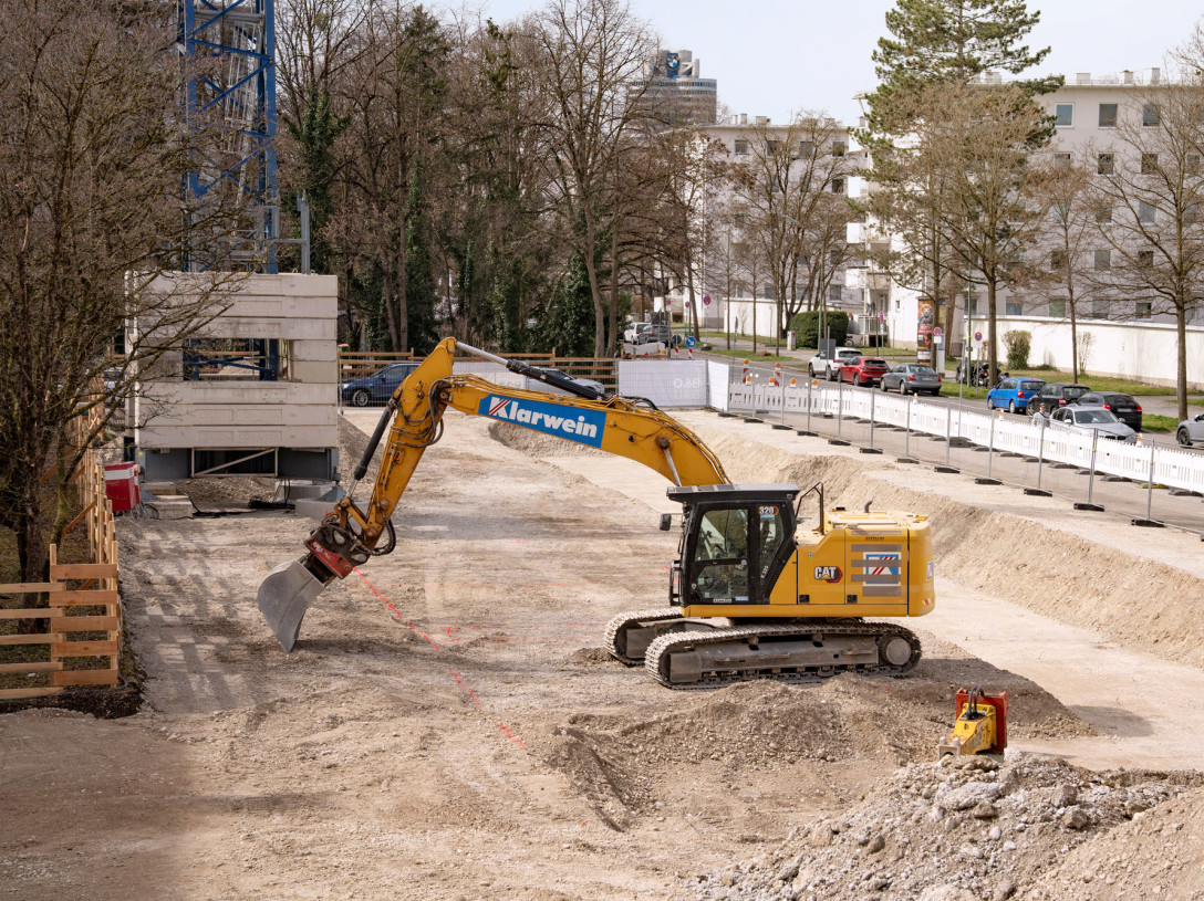 Ein Bagger gräbt in einer Baugrube. Rechts davon ist eine Straße zu erkennen und weitere Wohnhäuser.