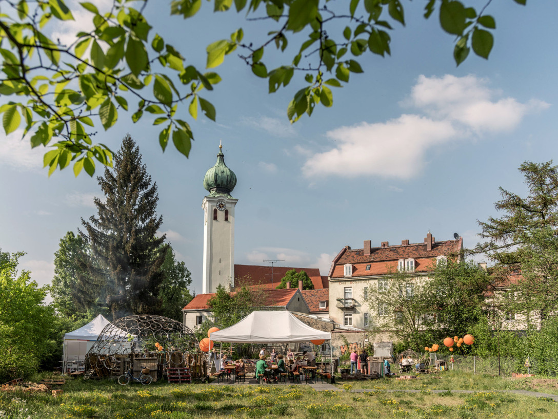 Blick auf einen wilden Garten in dem ein Pavillon und Bierbänke stehen auf denen Menschen sitzen. Im Hintergrund ist ein Kirchturm und ein Wohnhaus.