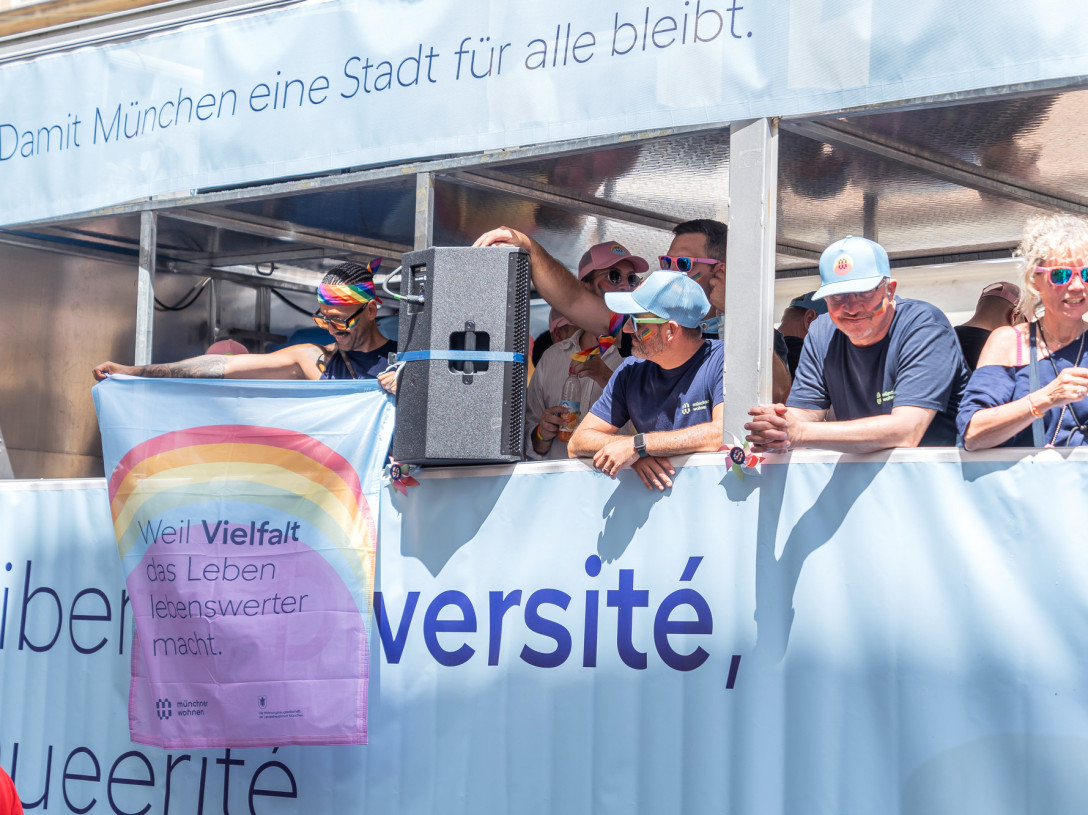 Mehrere Menschen stehen auf der Plattform eines geschmückten Trucks. Einer hält eine Fahne auf der steht "Weil Vielfalt das Leben lebenswerter macht.".