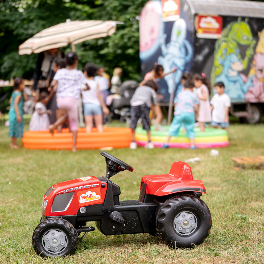 Im Vordergrund steht ein Bobby Car Auto, im Hintergrund erkennt man Kinder die spielen und in ein Planschbecken hüpfen.