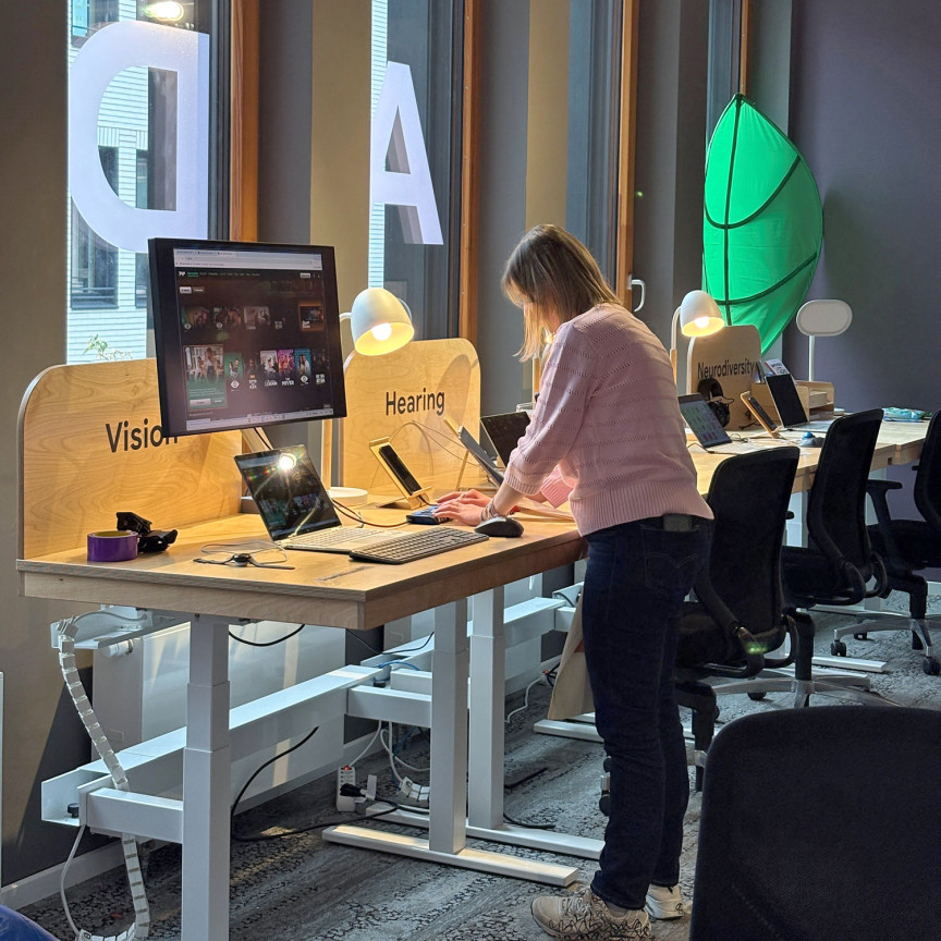 Eine junge Frau steht vor einem Tisch auf dem mehrere technische Geräte wie Laptop, Bildschirm und Smartphone angeordnet sind.