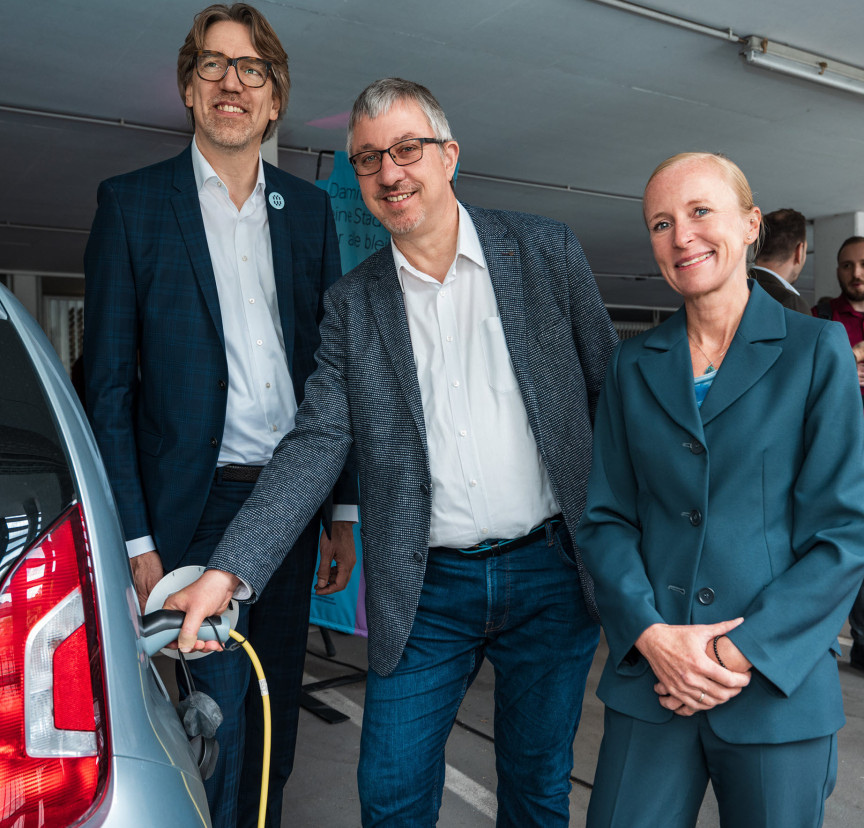 Gruppenbild mit 2 Männern und 1 Frau. Ein Mann hält den Ladestecker ans Auto. Auf dem Auto ist das bunte Münchner-Wohnen-Logo zu sehen.
