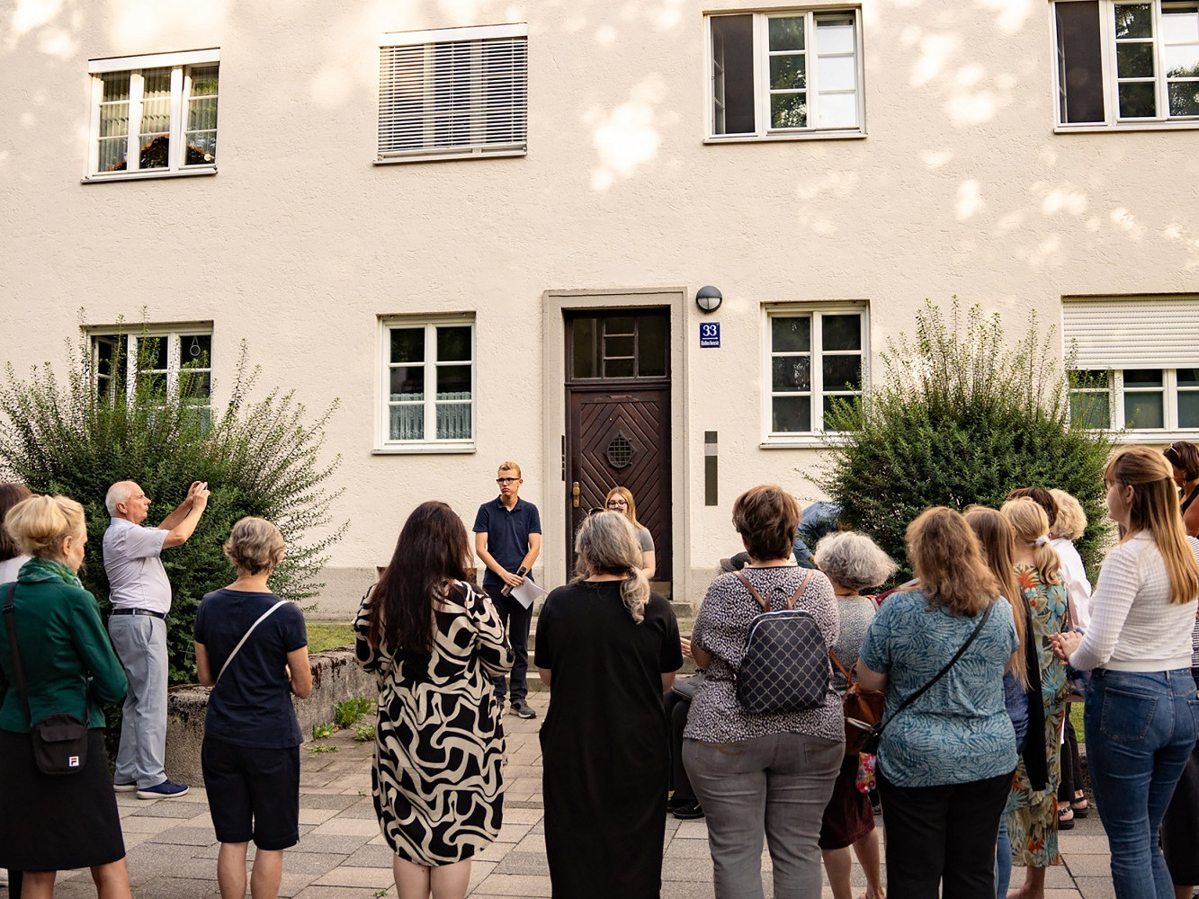 Eine Gruppe von Menschen steht vor einem Haus an dessen EIngangstür eine Plakette als Erinnerungszeichen angebracht wurde. Vor der Gruppe stehen zwei Menschen und sprechen.