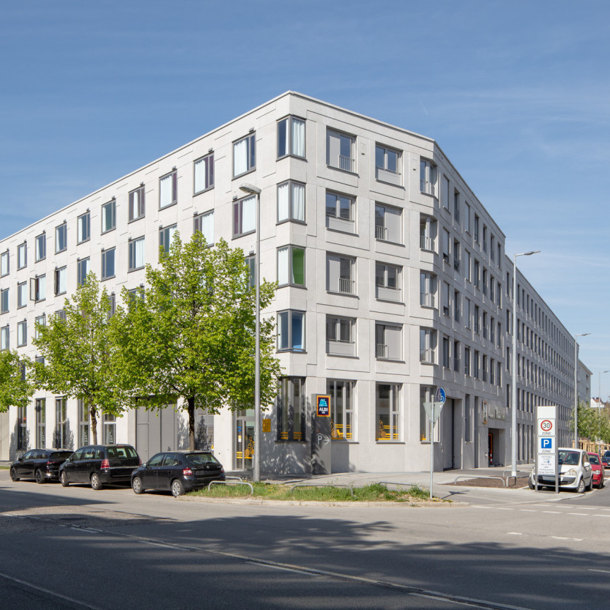 Blick auf ein mehrstöckiges modernes Wohngebäude, das an einer Straßenkreuzung steht. Davor viel Straßen- und Radverkehr sowie Menschen.